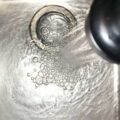 Image shows a stainless steel kitchen sink with a shower head mixer tap pouring water into the sink. The plug is closed or blocked as water is collecting in the sink.