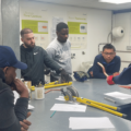 Image shows a group of plumbing students being taught about pipe bending. They are gathered around a workbench and the instructor is holding a piece of copper pipe. There is a pipe-bending tool on the work bench,