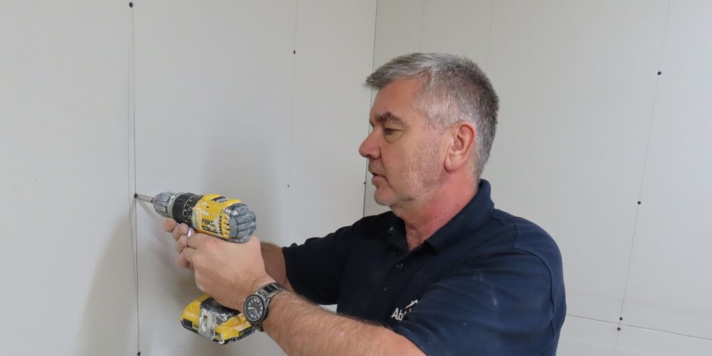 A person is using a cordless screwdriver to screw plasterboards onto a wall. They are dry-lining an internal wall