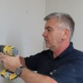 A person is using a cordless screwdriver to screw plasterboards onto a wall. They are dry-lining an internal wall