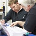 Two men are reviewing documents together in a workshop or training environment. One man, with short light-colored hair, points at the papers while the other man, with short brown hair, looks closely and holds a pen, appearing to take notes or discuss details. Tools and equipment can be seen in the background, suggesting a technical or vocational setting, possibly related to gas training or certification.