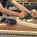 Image shows a workbench where someone is using a plane to shave wood. There are three pieces of wood on the bench indicating it is a workshop or construction setting.