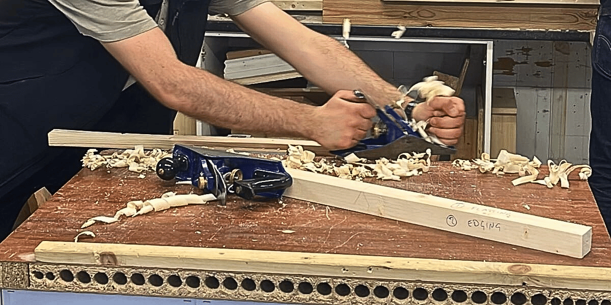 Image shows a workbench where someone is using a plane to shave wood. There are three pieces of wood on the bench indicating it is a workshop or construction setting.