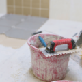 Image shows a floor, possibly a bathroom or kitchen, as tile are being put on the floor, In the foreground there is a bucket, with a grouting tool, dustpan and brush and another larger bucket. In the background, a person is kneeling on the floor laying tiles or positioning tile spacers.