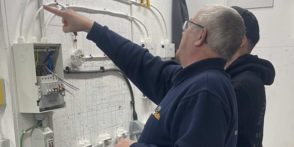 Image shows an instructor and a student working on electrical installation. On the wall in front of them is a series of wires, switches, and sockets. The instructor is pointing to something.